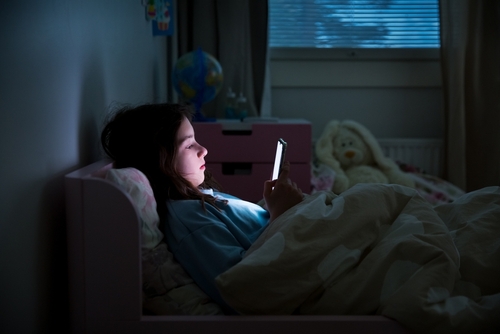 A young girl lying in bed at night, illuminated by the glow of a smartphone screen while using the device under a blanket.